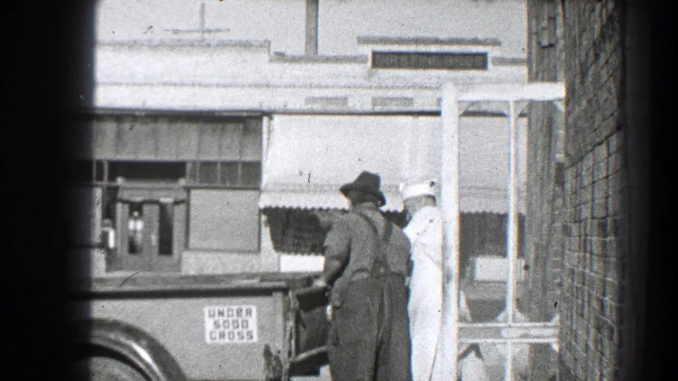 Two men are visible near the truck rear, one in overalls and one in white clothing, with storefront signage and a brick wall in the frame.