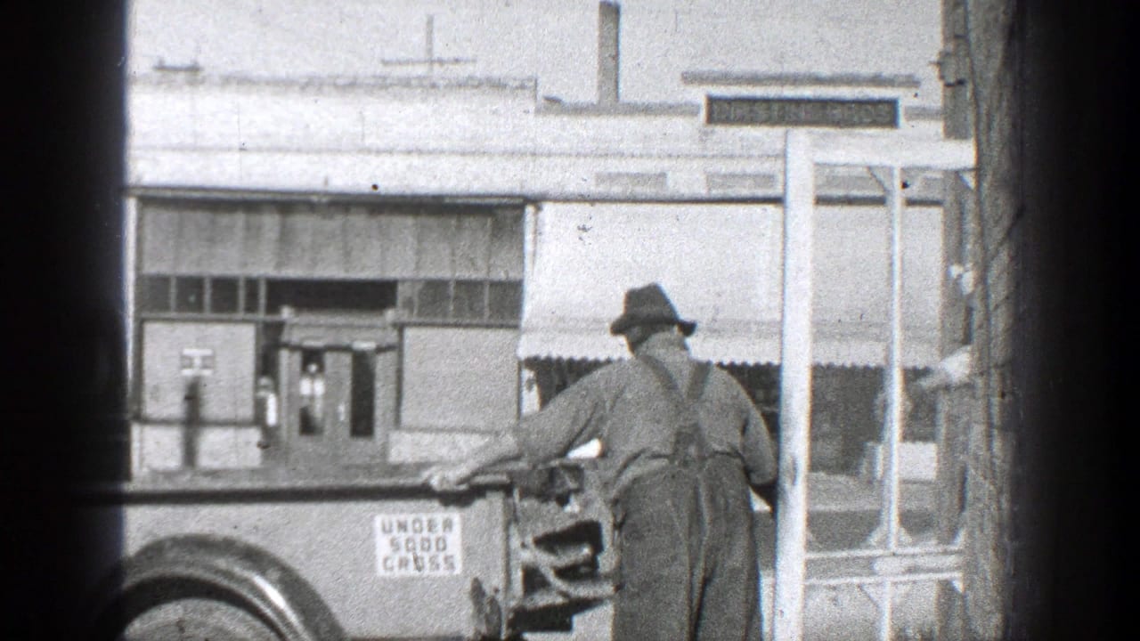 Man in hat and overalls stands beside a vintage truck in front of a storefront.