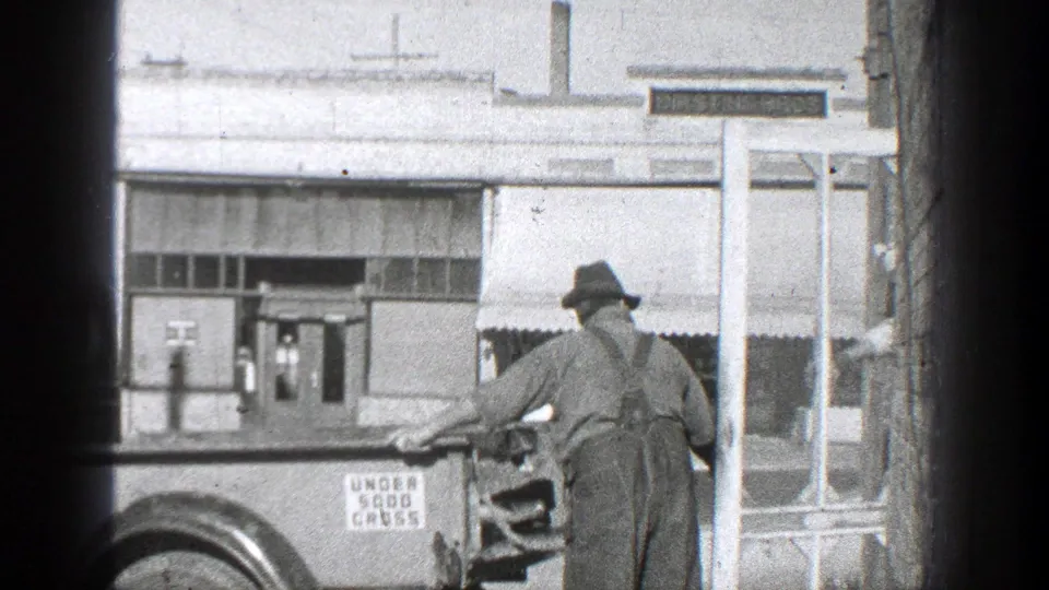 In 1936 California home-movie footage, a man in hat and work clothes stands by a parked vintage truck near a commercial building.