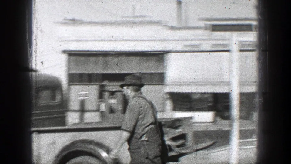A man in work clothing and hat is seen beside a vintage truck parked along a storefront in the stated California 1936 setting.