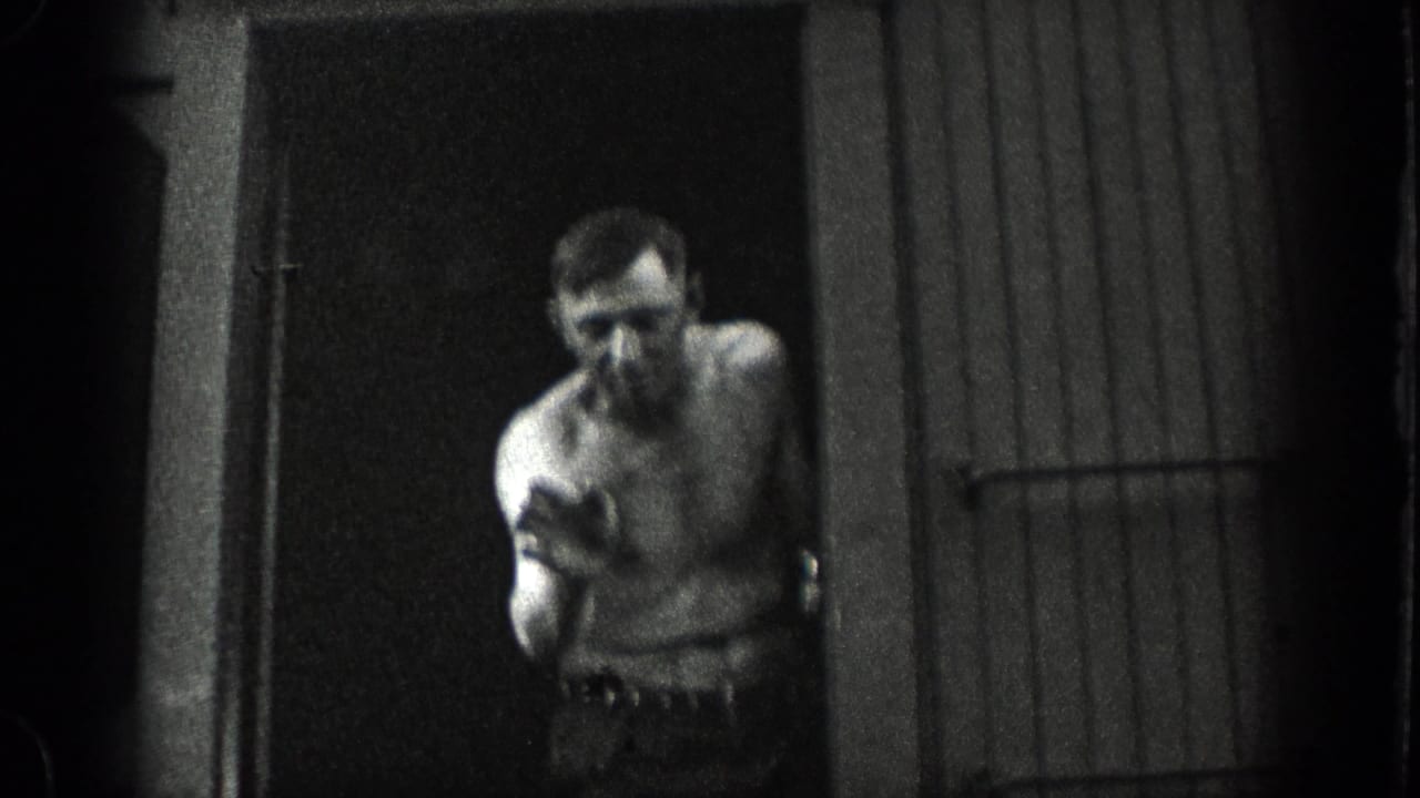 1936 California home-movie frame showing a shirtless man near a doorway or shed entrance, with soft focus and heavy shadow.