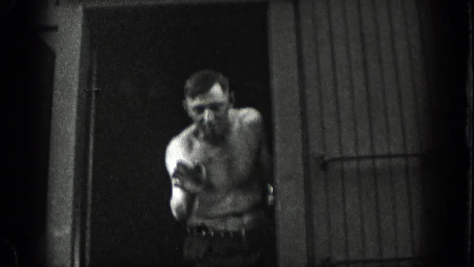 1936 California home-movie frame showing a shirtless man near a doorway or shed entrance, with soft focus and heavy shadow.