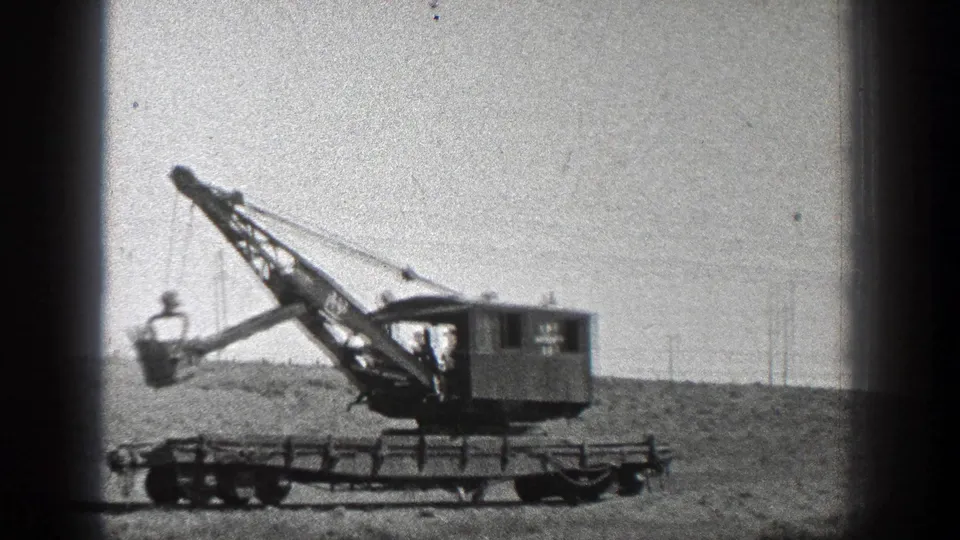 1936 California 8mm frame of a wheeled industrial crane with a boom and bucket against a bright open sky.
