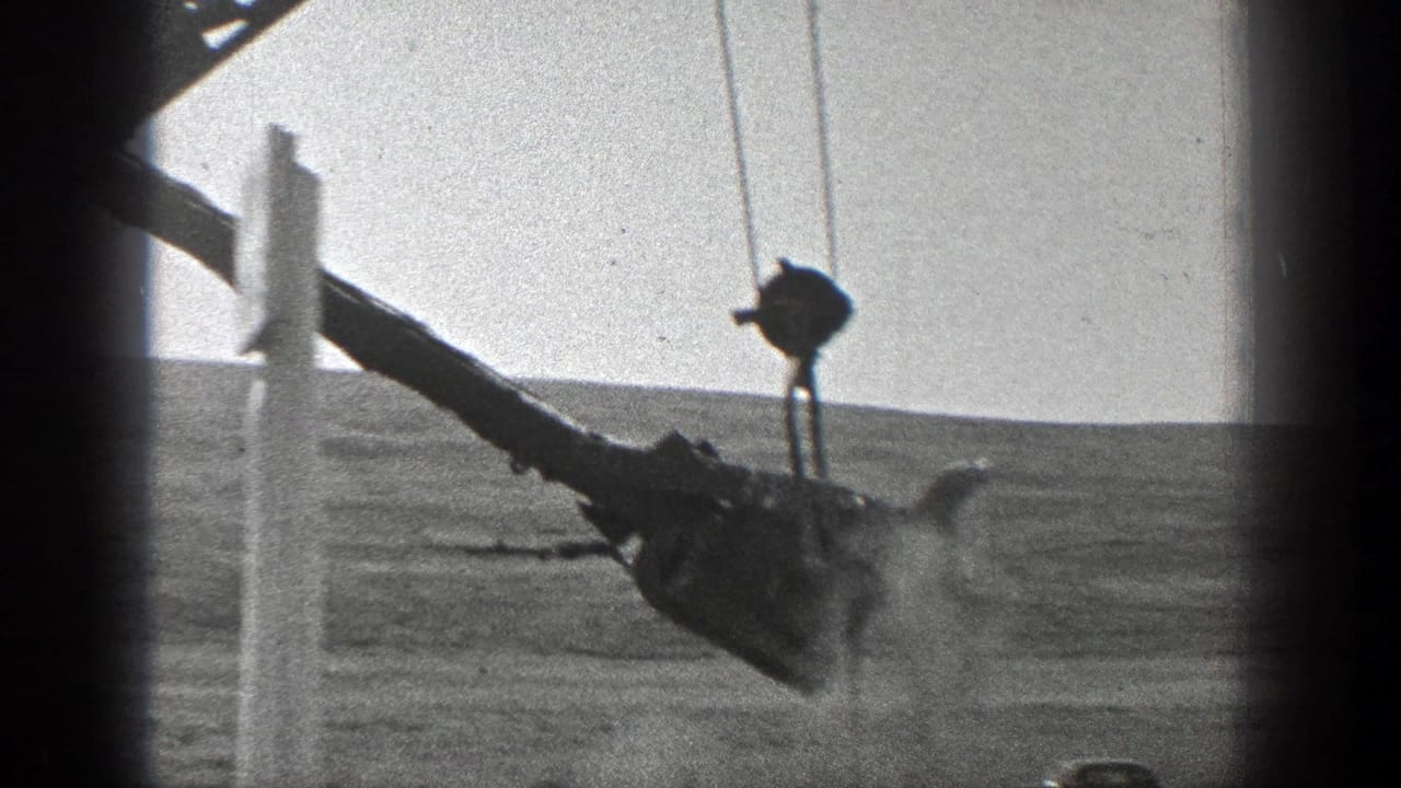 Crane boom lifts or lowers a bucket near a vertical post over dusty ground.