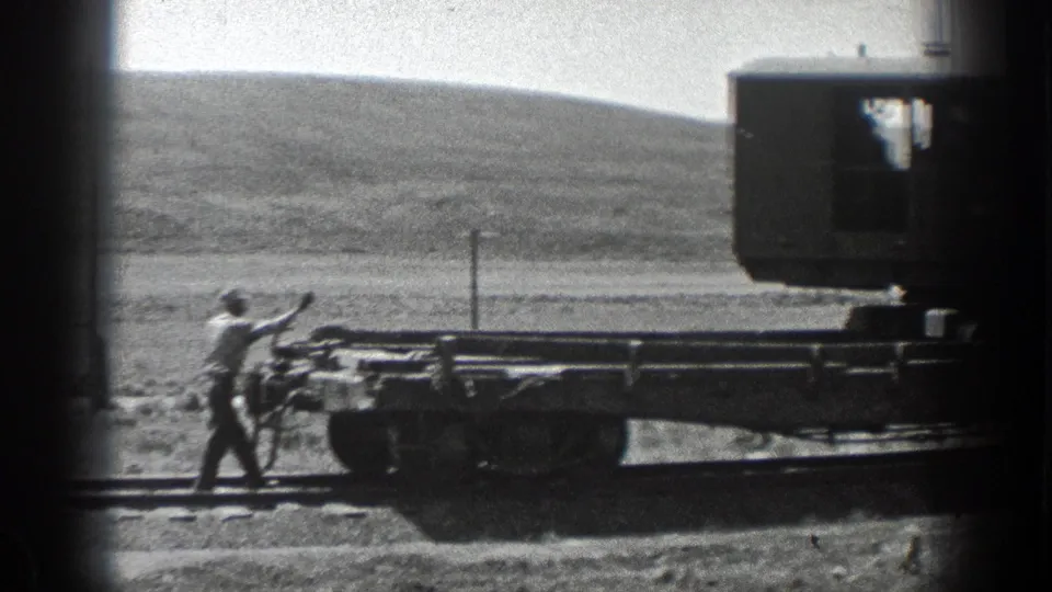 1936 California 8mm frame showing a worker reaching toward the rear of a flat railway car on rural railroad tracks.