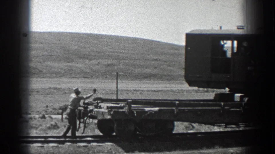 Worker in 1930s California footage stands at the rear of a flat railway car while dark rail equipment fills the right side of the frame.