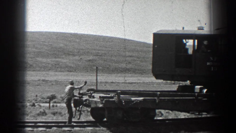 1936 California rail scene with a worker beside a flat car, one arm raised toward the brake or coupling area.