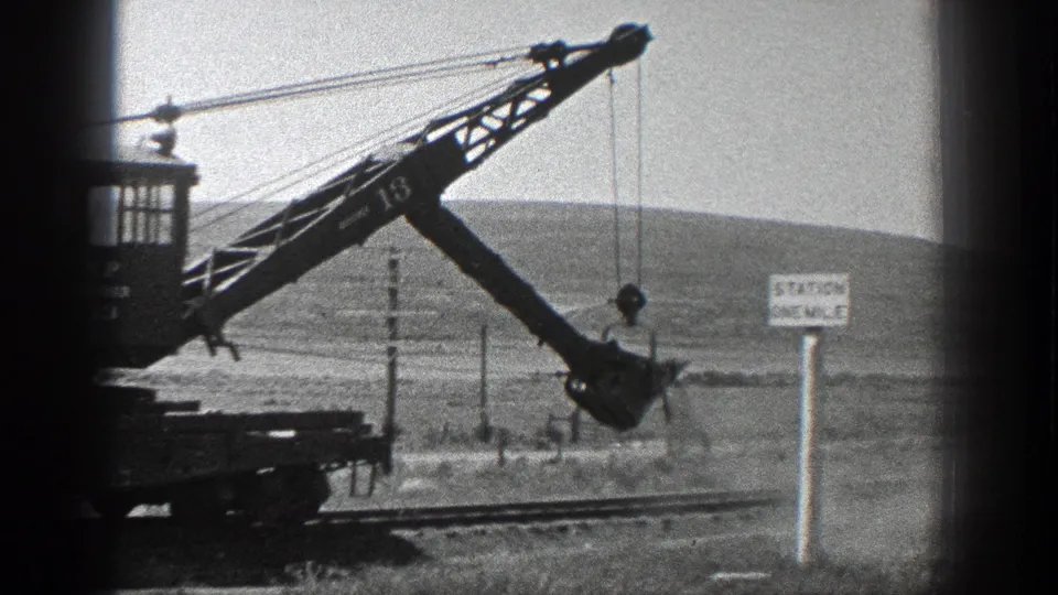 1930s rural rail-work view with crane boom extended, bucket suspended, track below, and the Station One Mile sign at right.