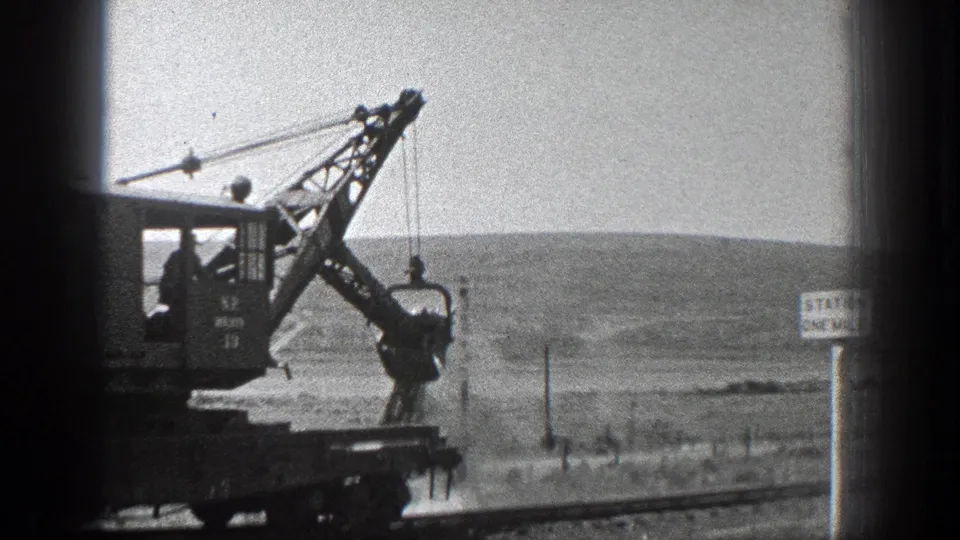 Black-and-white 1936 California frame showing crane rail equipment near tracks, with open land and a small sign in the background.
