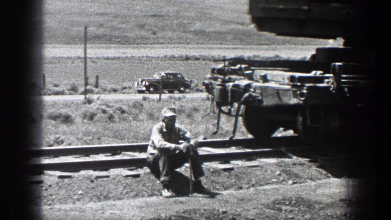Man sits near railroad tracks beside rail equipment and a distant car.