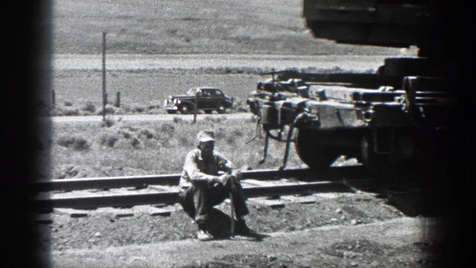 A man in work clothes sits by railroad tracks in 1936 California, with rail machinery at right and a vintage car beyond the line.