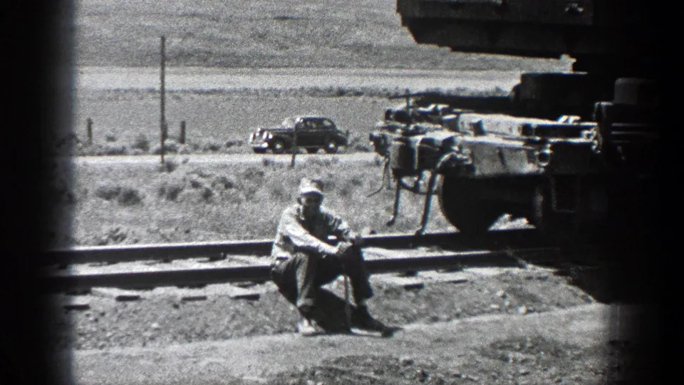 Black-and-white 8mm view of a man seated beside railroad tracks in California, with machinery and a car visible behind him.