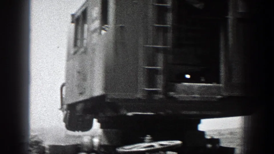 Rear portion of a large crane cab in black-and-white 8mm footage, with ladder, undercarriage, and mechanical parts visible.