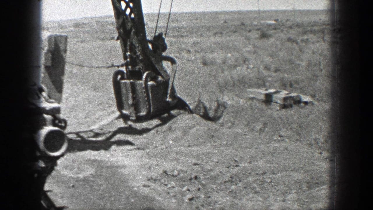 Suspended crane bucket and cables over sandy ground with shoreline in the distance.