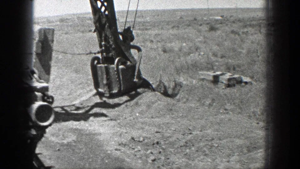 1936 California frame showing a crane bucket hanging from cables over sand, with open coastal terrain and waterline beyond.