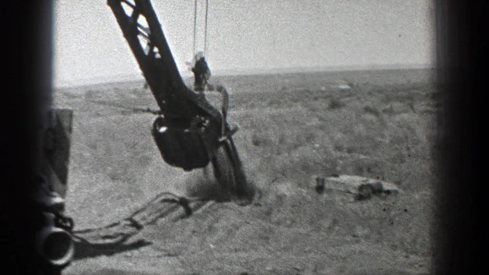 1936 California 8mm frame showing the crane boom, lowered bucket, cables, sandy terrain, and a small worker figure near the machinery.