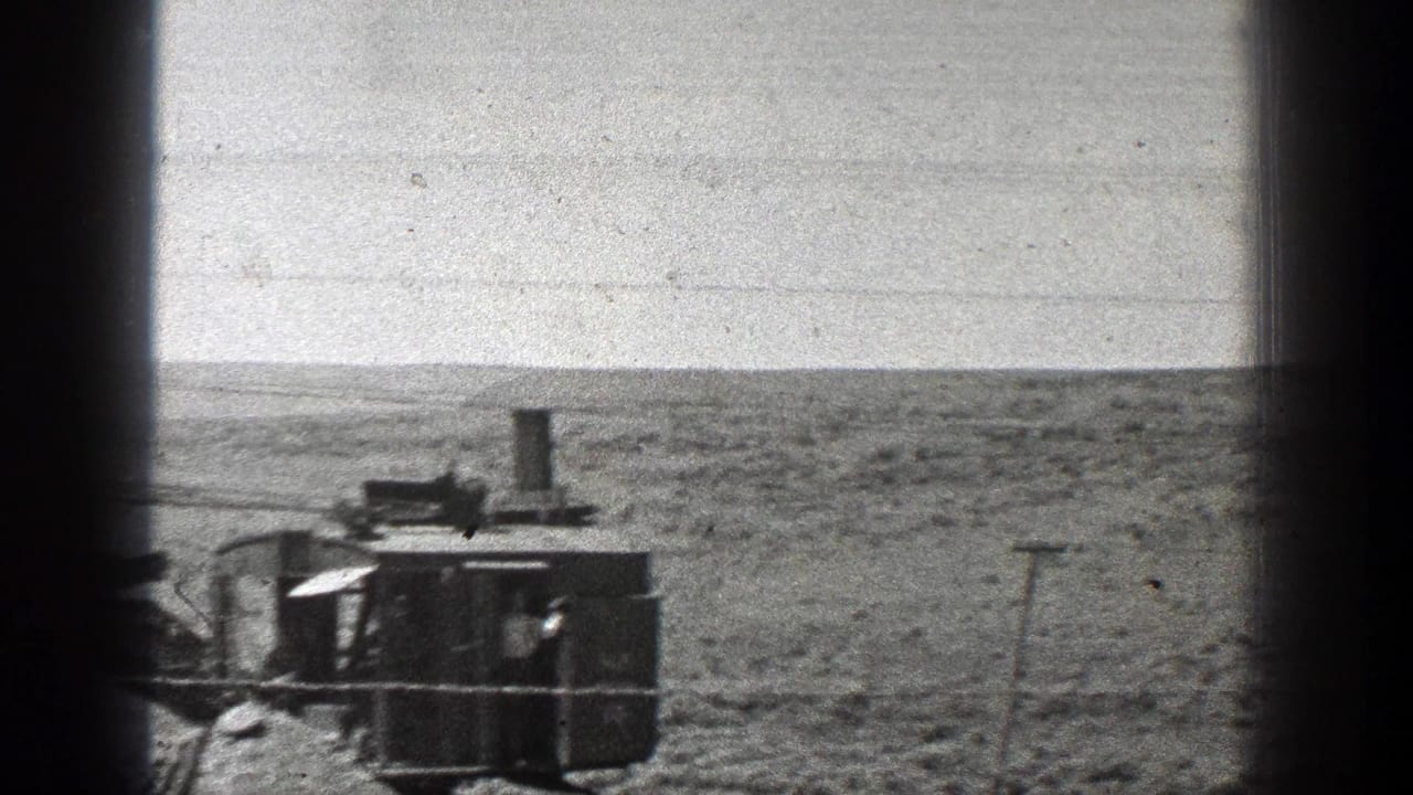 Steam-powered earth mover seen through dark window edges in open desert terrain.