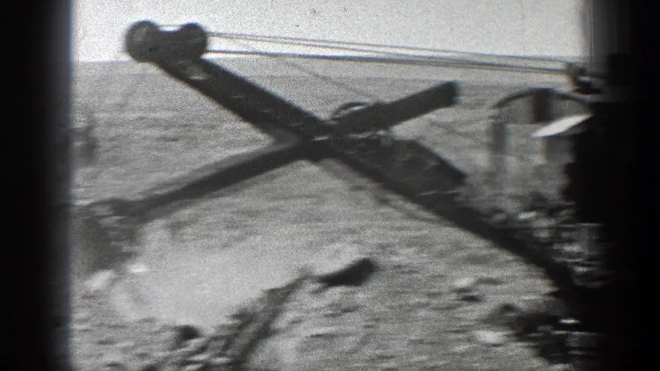 Motion-blurred 8mm frame showing the earth mover's cable boom or scoop mechanism crossing an arid California work site.