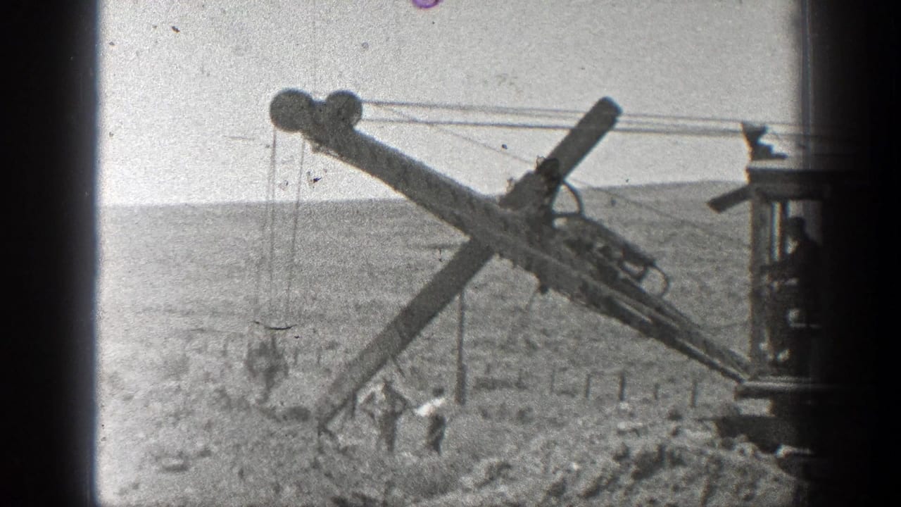 Drilling machine boom over dusty ground with workers near the rail bed.