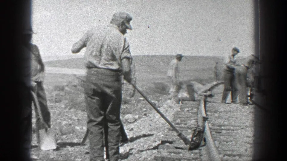 In 1936 California, adult male workers stand and shovel beside a newly laid railroad track in a dusty open landscape.