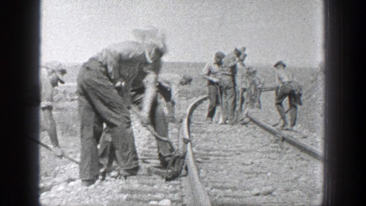 Railroad workers bend over tools and track in a bright outdoor setting.
