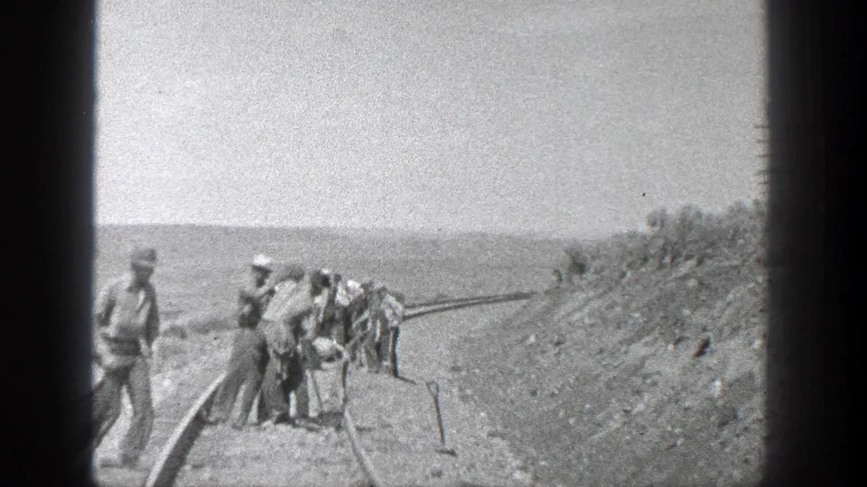 A crew in work clothes and hats appears to dig and align railroad track in a flat California landscape, with rails extending through the scene.