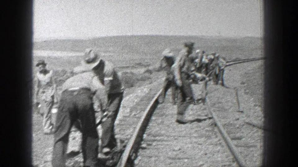 Railroad construction workers in 1930s attire work with shovels and rail equipment as the track curves across an open California setting.