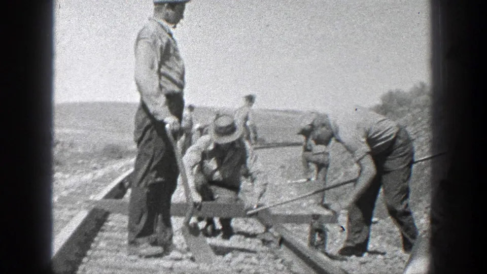 A closer view shows several workers using hand tools beside railroad rails, with one man standing near the track and others bent over their tasks.