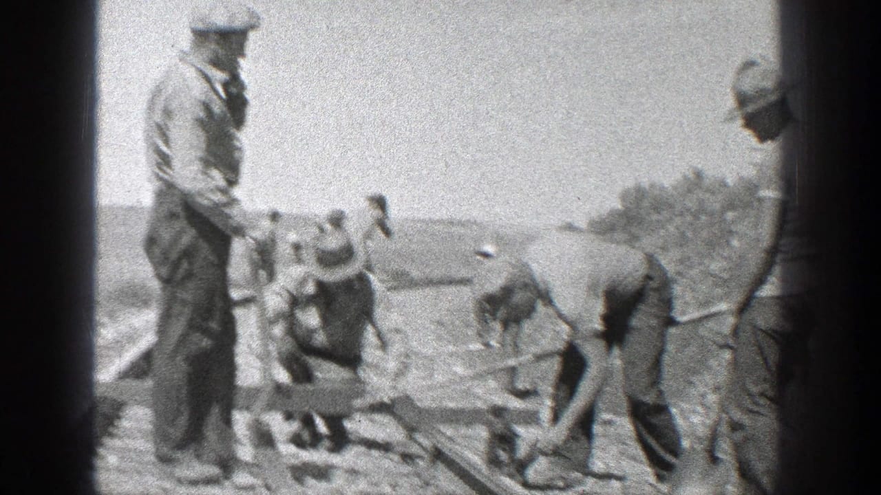 Men in hats work beside railroad tracks with tools in an open landscape.