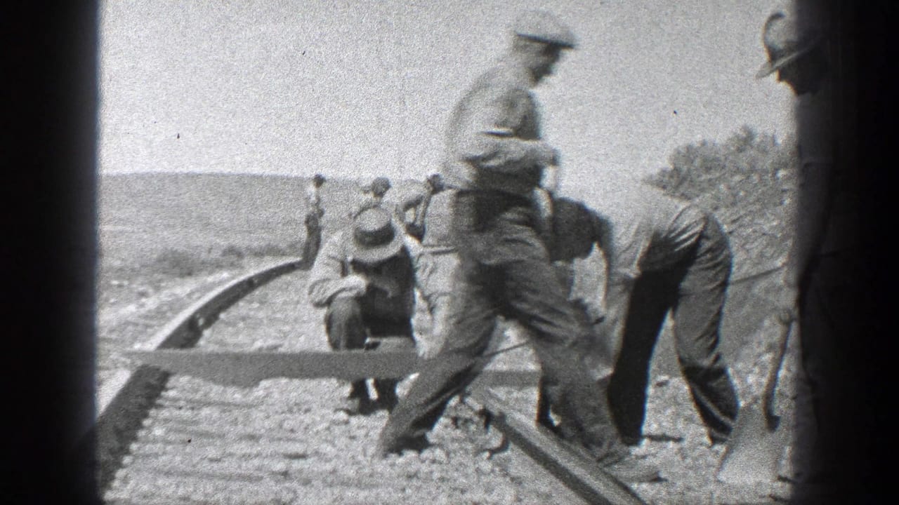 Workers stand and crouch along railroad tracks while handling a long timber or rail component.