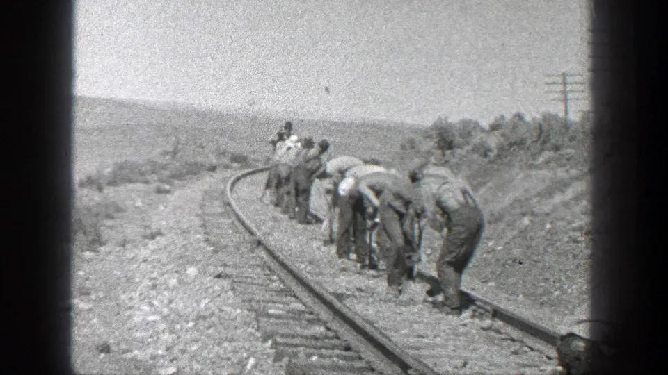 Men in work clothes bend over a curved railroad track in rural California, with open hills, gravel ballast, and utility poles visible in the distance.