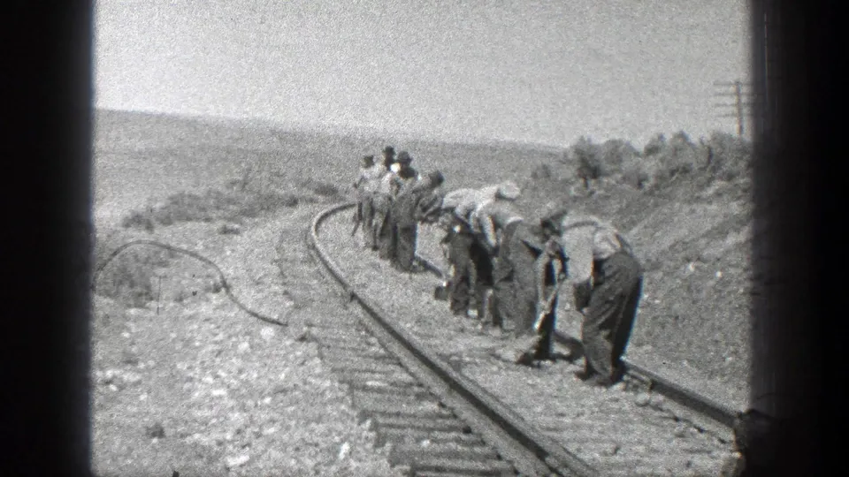 A closer view of men bent over railroad ties and rails, appearing to use hand tools on the track in the arid rural California setting.
