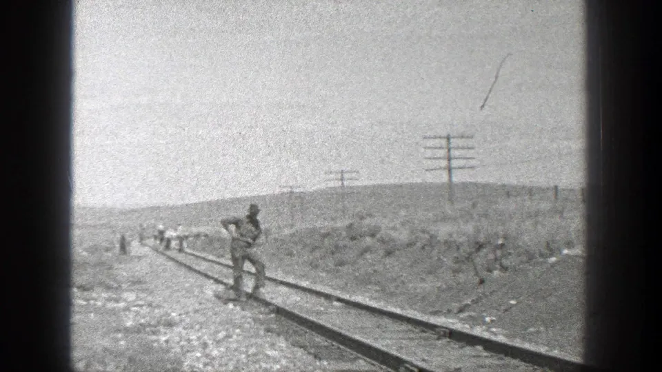 Wide shot of a rural rail line with several workers in the distance and one man nearer the track, framed by open terrain and utility poles.