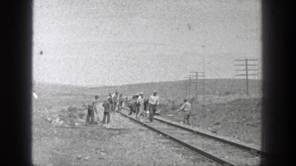Workers in period clothing cluster along railroad tracks in rural California, with poles and low terrain visible behind them.
