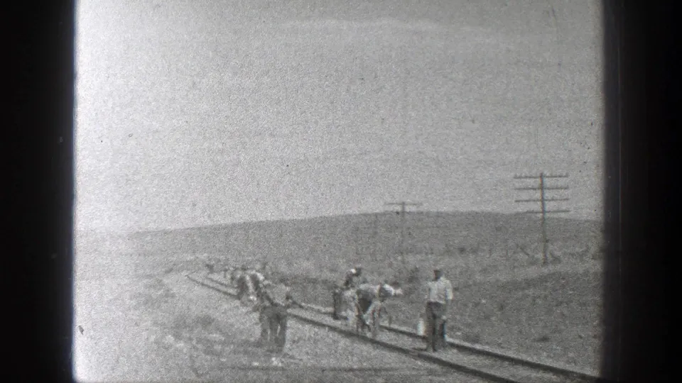 Distant workers appear to maintain or walk along a railway line in a sparse California desert setting.