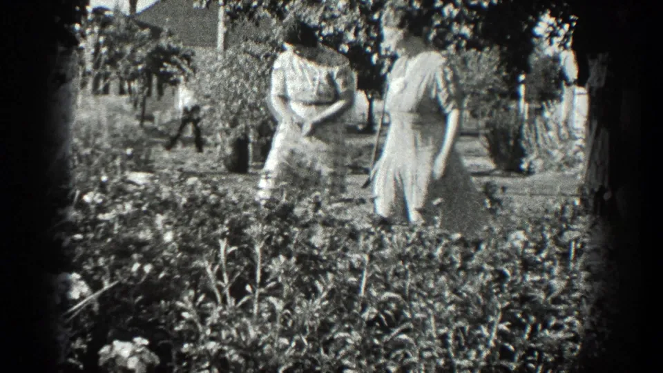 Two adult women in dresses pause near flower beds and trees in 1936 California garden footage.