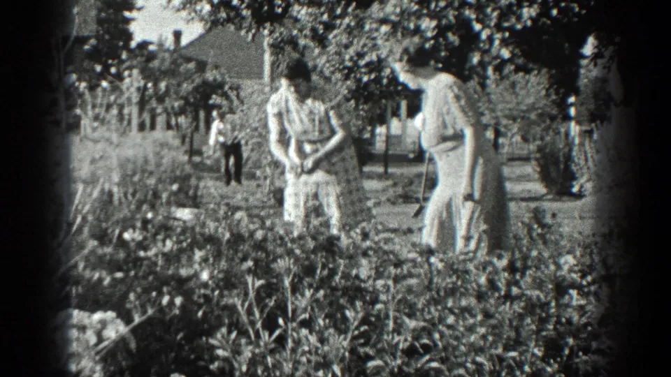 1936 California home-movie frame of women in light dresses beside a lush garden, with a house and distant person in the background.