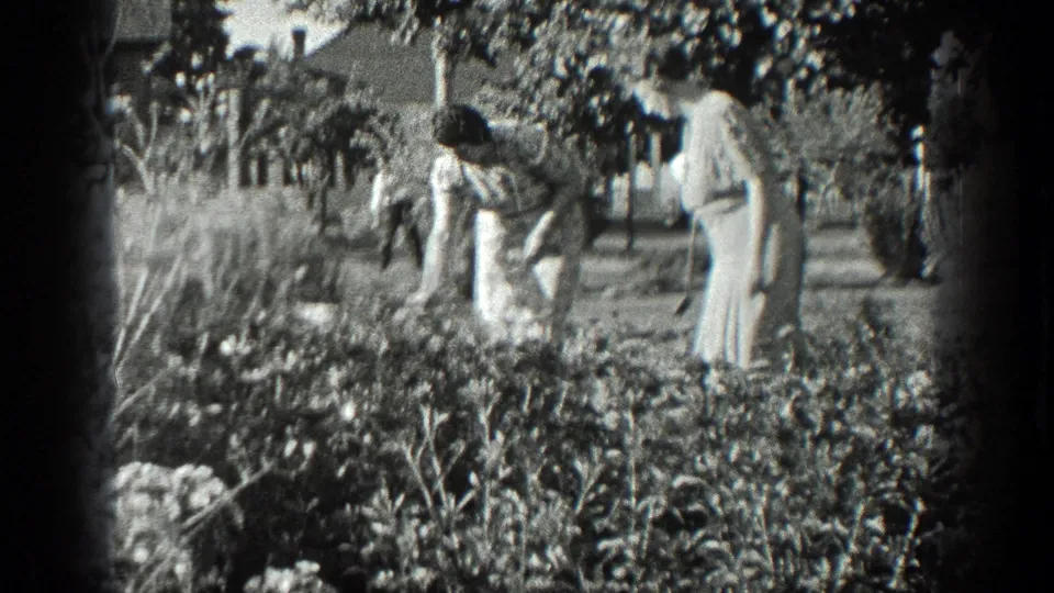 Black-and-white 8mm frame from 1936 California showing one woman reaching into a flowerbed as another watches nearby.