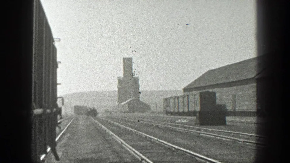 Black-and-white 8mm footage shows rail lines, freight cars, and a smokestack from a moving train perspective.