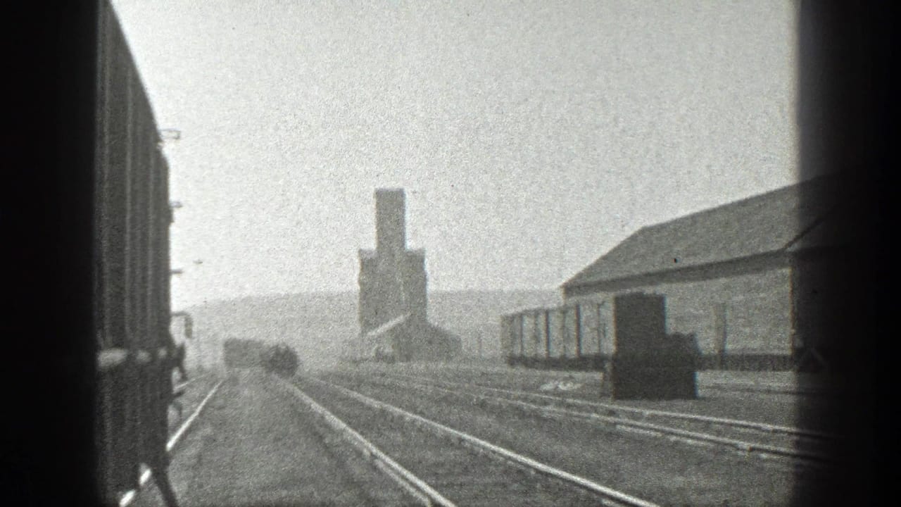 Rail tracks seen from a train window beside freight cars and an industrial building.