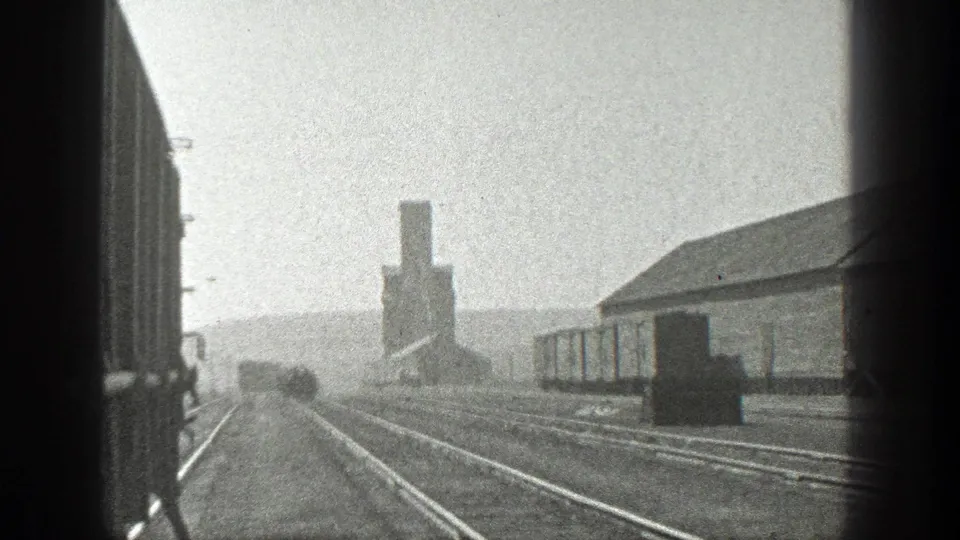 1936 California 8mm view from a moving train, looking along rail tracks toward freight cars, an industrial building, and a distant silo-like structure.