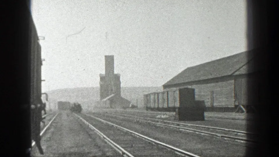 1936 California home-movie frame of a rail yard from a moving train, with multiple tracks, freight cars, and a low industrial building.