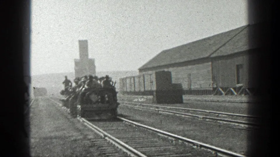 1936 California 8mm frame of men riding a small rail vehicle past freight cars, tracks, and a low industrial building.