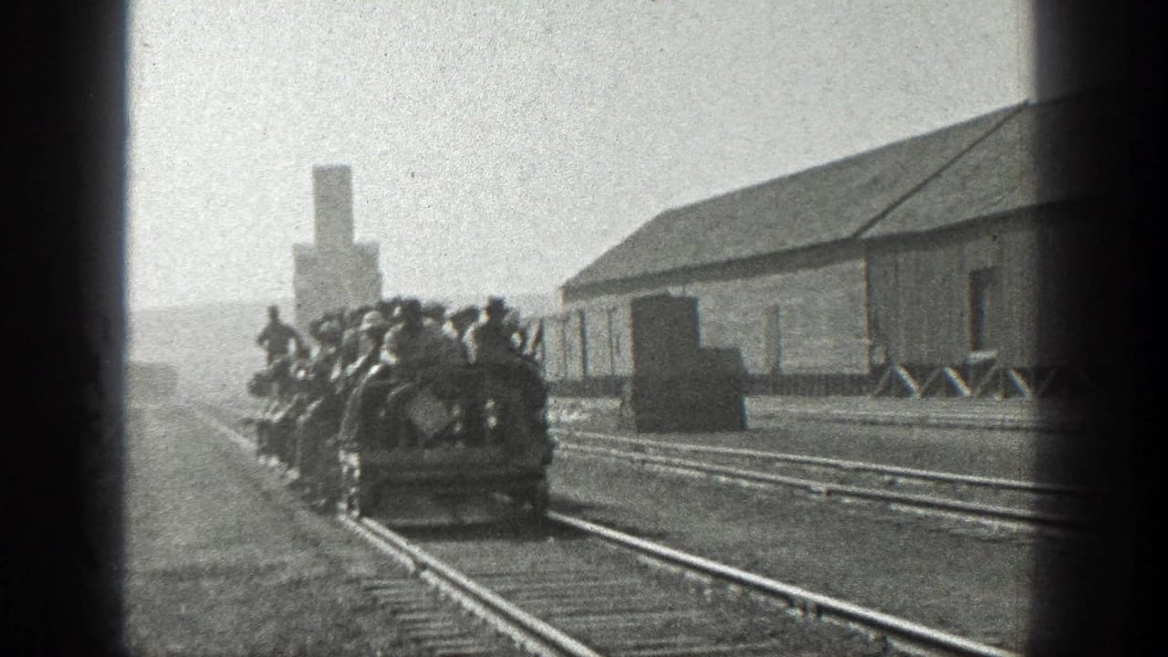 1936 California home-movie frame of crowded open rail cars with adult men riding past a wooden industrial building and tracks.