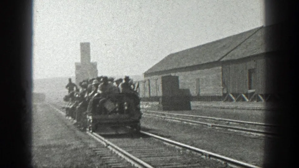 1936 California home-movie frame of crowded open rail cars with adult men riding past a wooden industrial building and tracks.