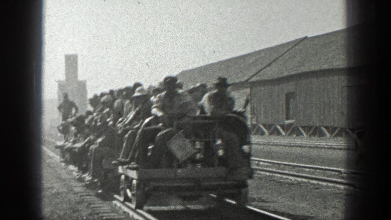 Crowded open rail cars move past the camera near a wooden building and tracks.