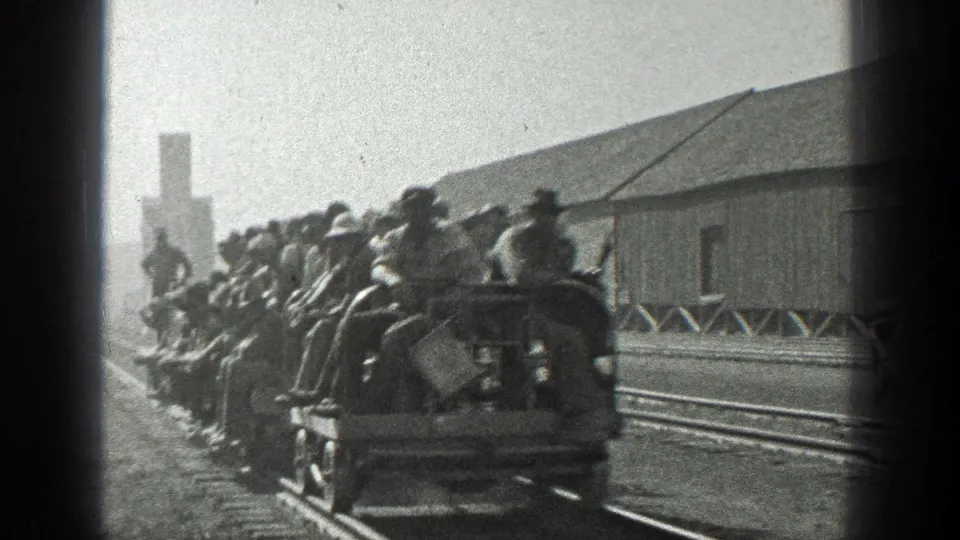Adult men in hats ride in crowded open freight cars in a 1936 California rail-yard scene with a wooden building nearby.