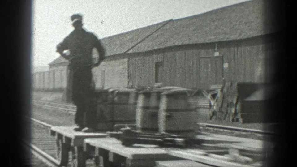 A man stands on a flat rail car carrying barrels in the 1936 California industrial rail scene, with tracks and a wooden building behind him.