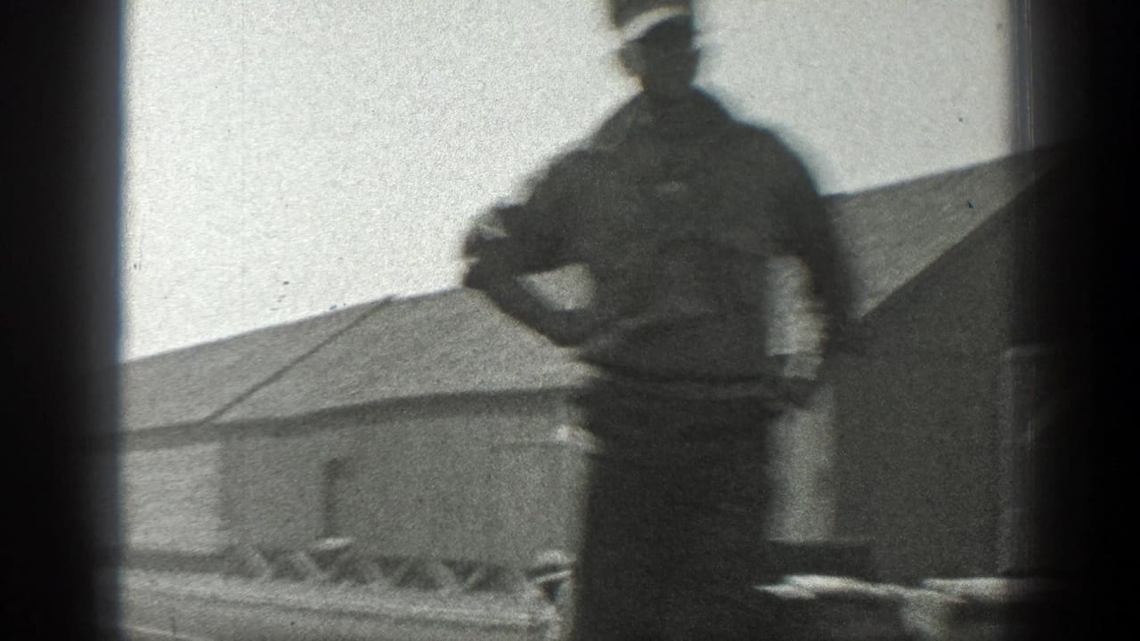 Uniformed man on a flatcar beside an industrial building, filmed from a moving train.
