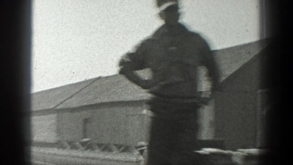 1936 California home-movie frame showing a uniformed man on a rail flatcar beside an industrial building as the train passes.